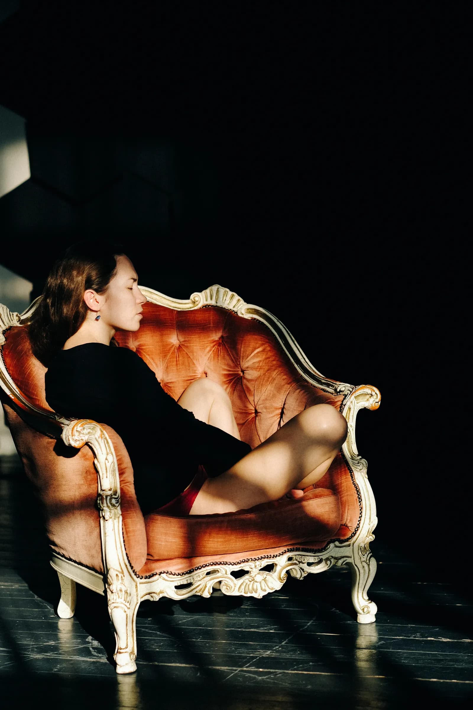 Woman asleep against the arm of a baroque armchair, hard light cutting the dark floor
