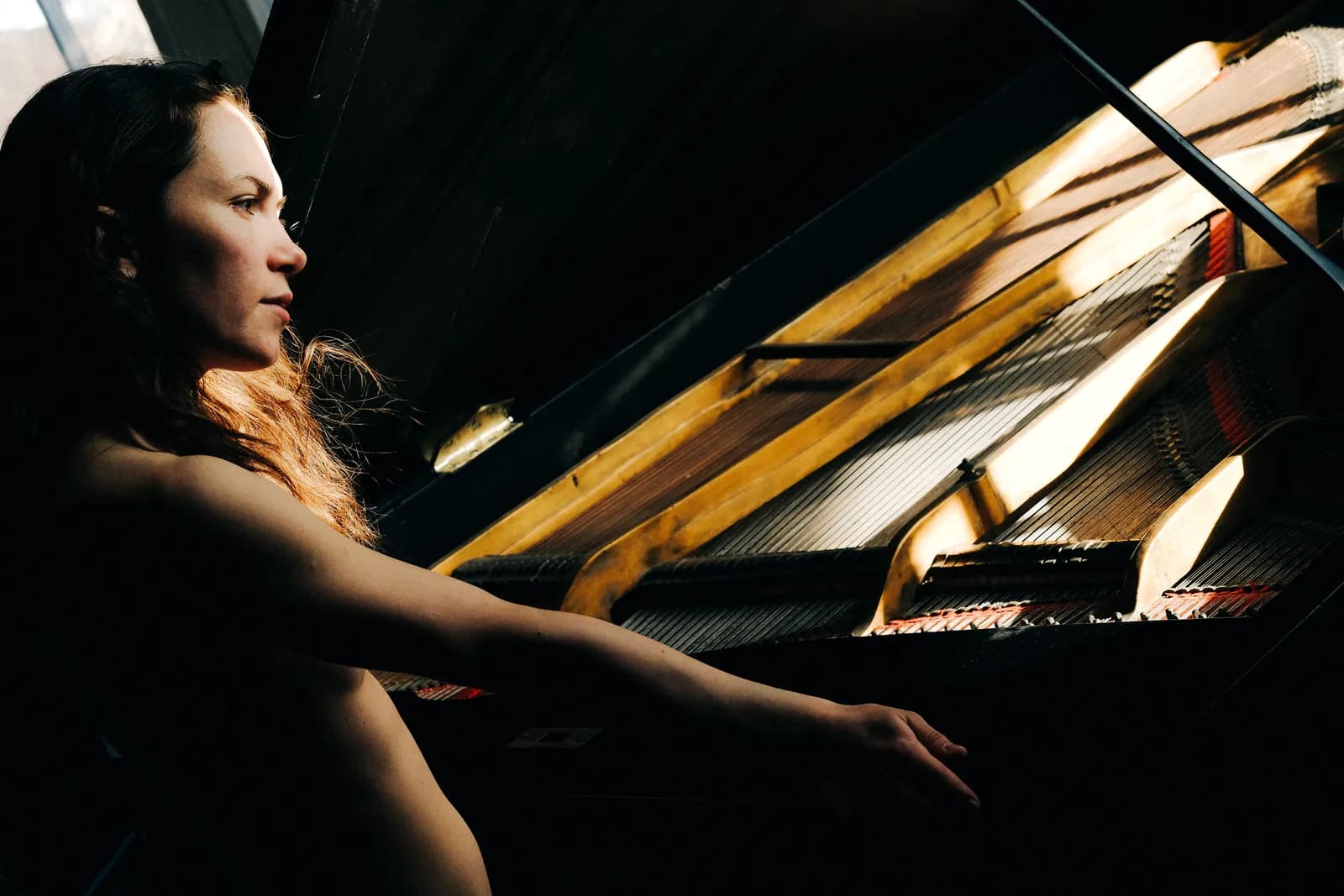 Nude woman in profile reaching across the open piano strings, hair lit by the window