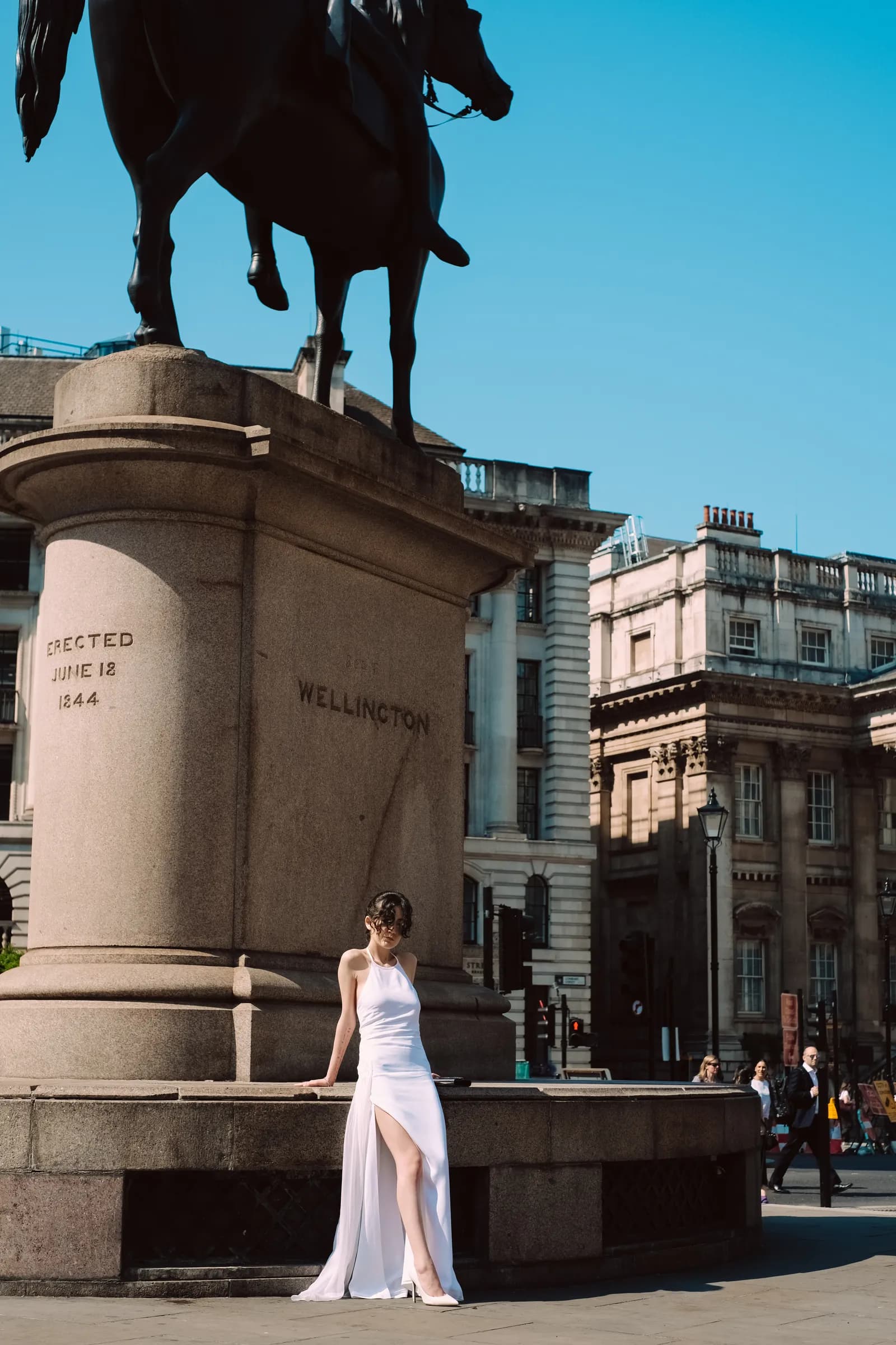 Woman in a white gown resting against the plinth of the Wellington statue at Bank, blue sky above