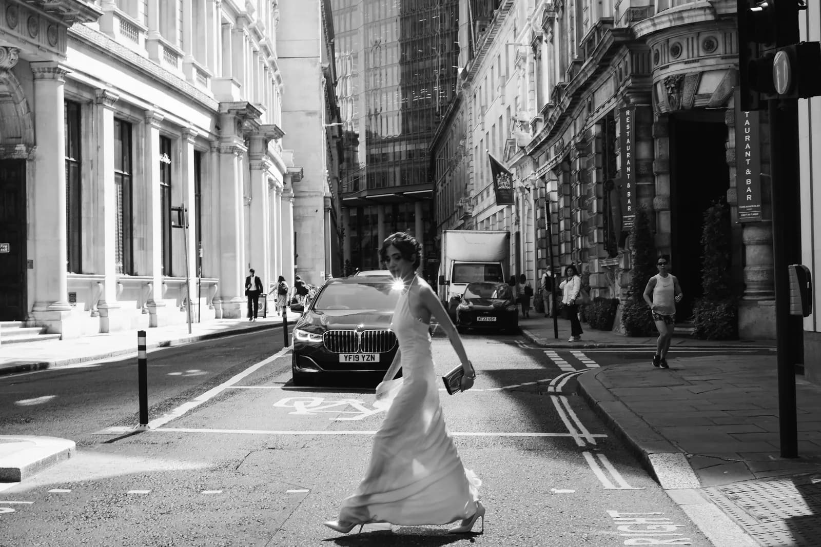 Black and white: woman in a white gown striding across a City street between black cabs