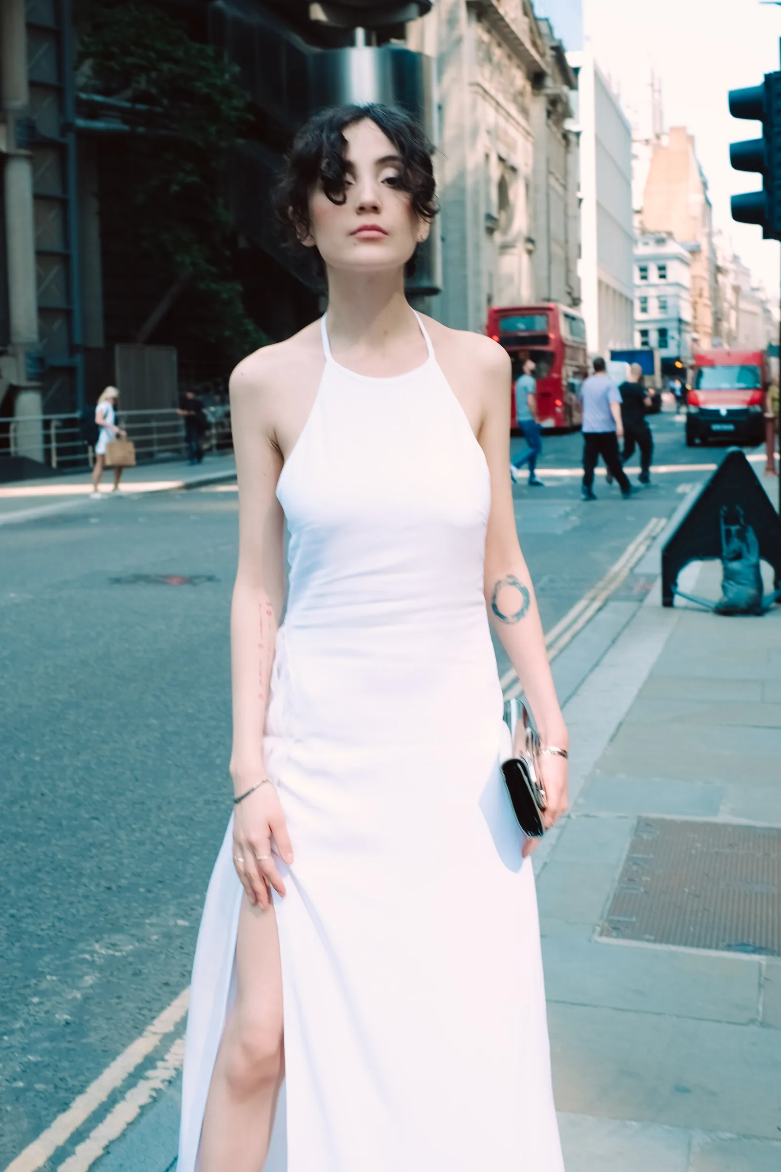 Woman in a white gown with a thigh-high slit on a City pavement, red buses down the street