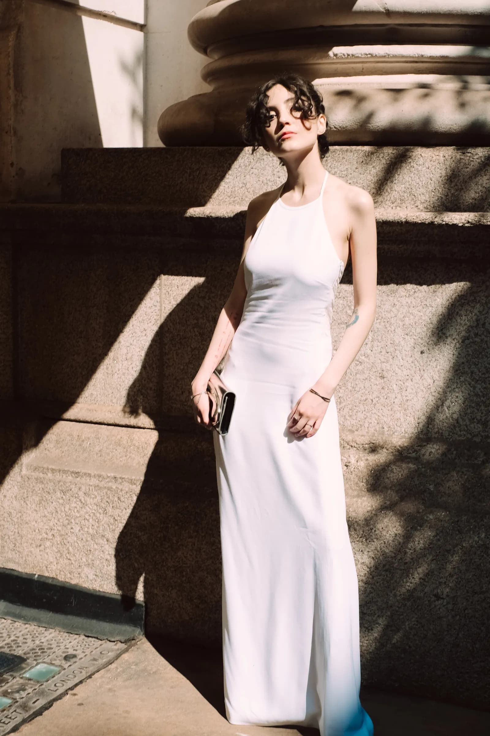 Woman in a long white halter dress in a shaft of sun against a monumental column