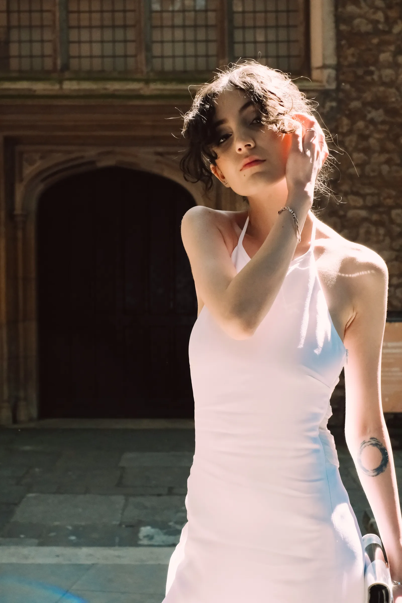 Woman in a white halter dress before a Tudor doorway, hand at her cheek, in hard sun