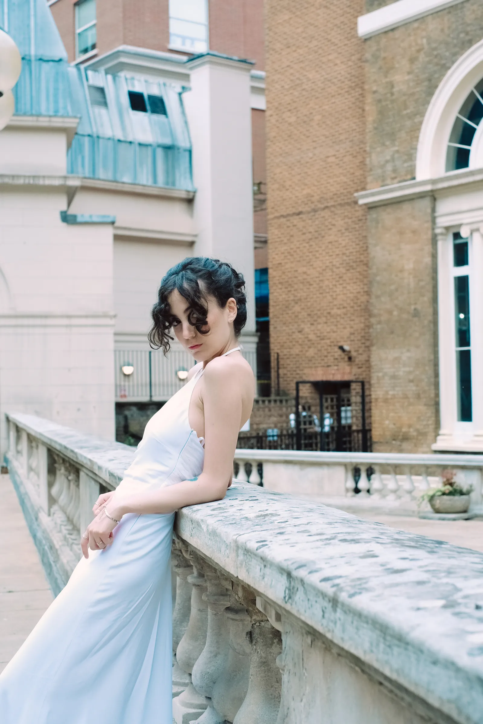 Woman in a pale slip dress leaning back on a stone balustrade, curls loose, Georgian brick behind