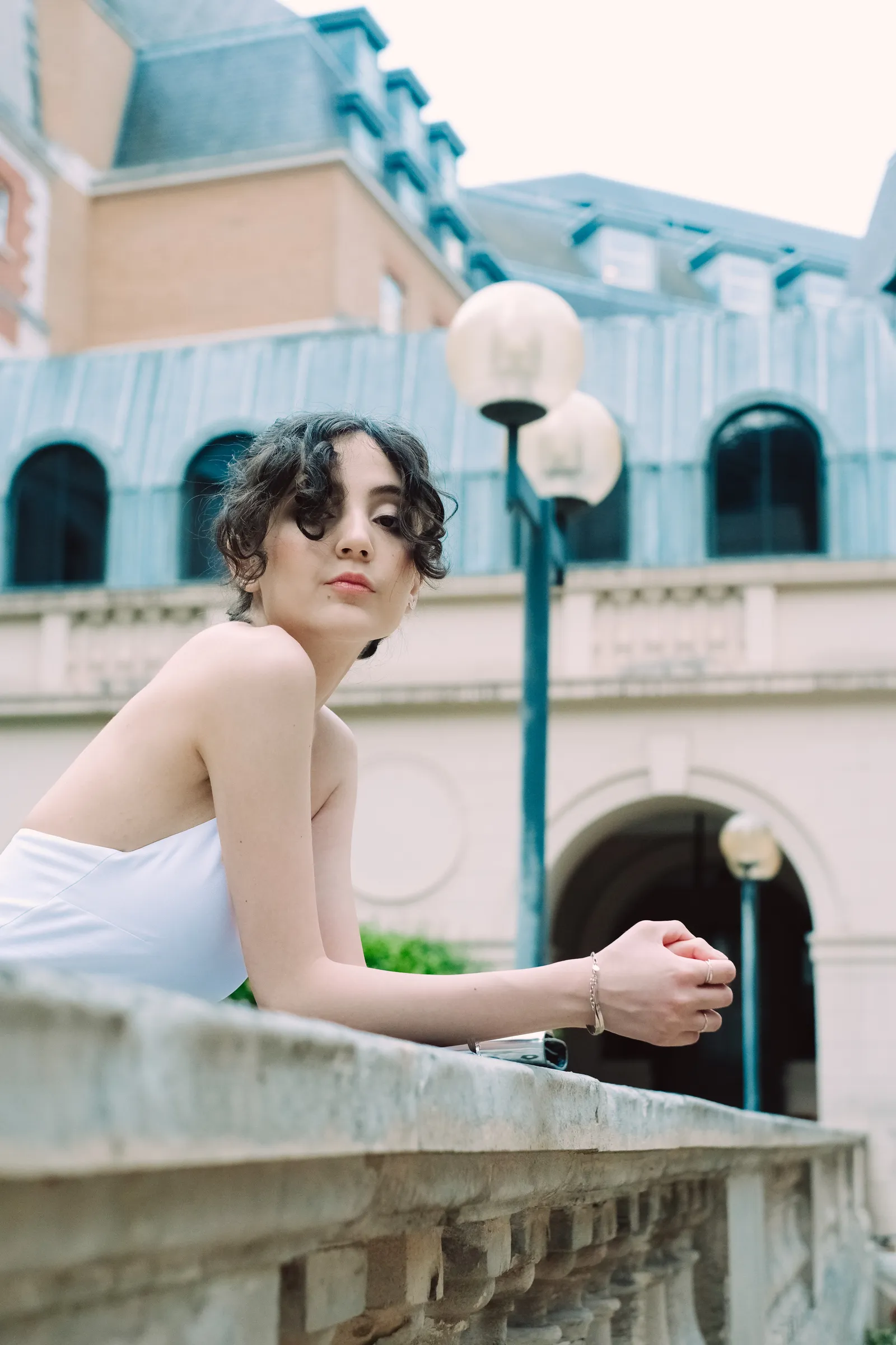 Woman in white leaning on a stone balustrade and looking back over her shoulder, grand facade behind