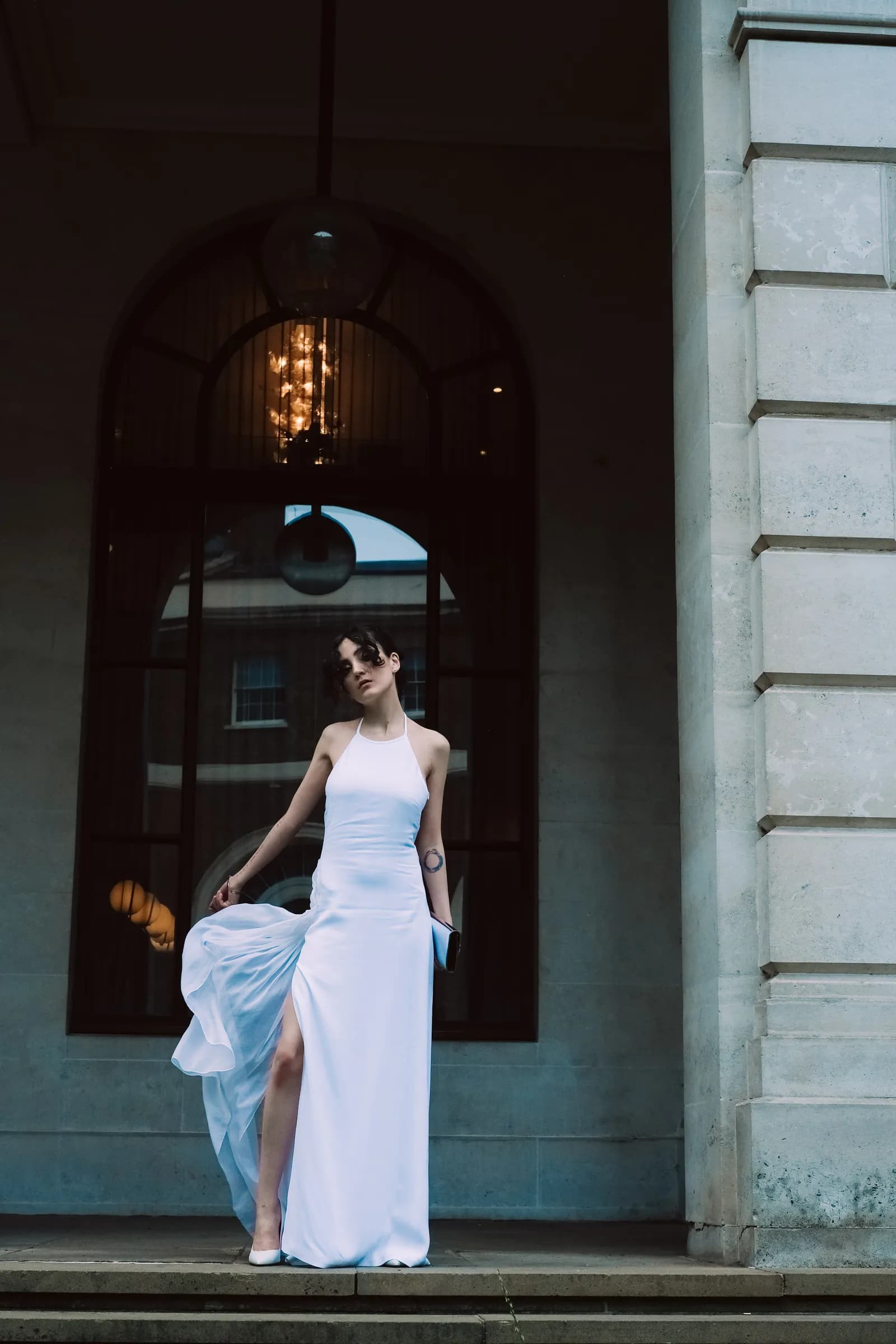 Woman on stone steps lifting the slit hem of her white gown, dark portico behind