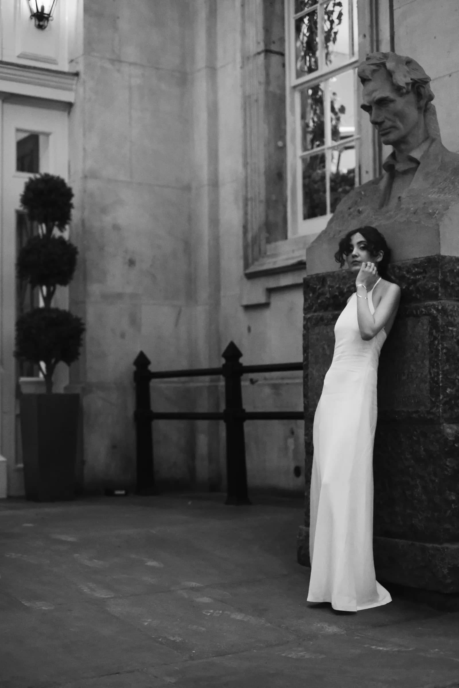 Black and white: woman in a white gown leaning against a stone bust outside a City portico