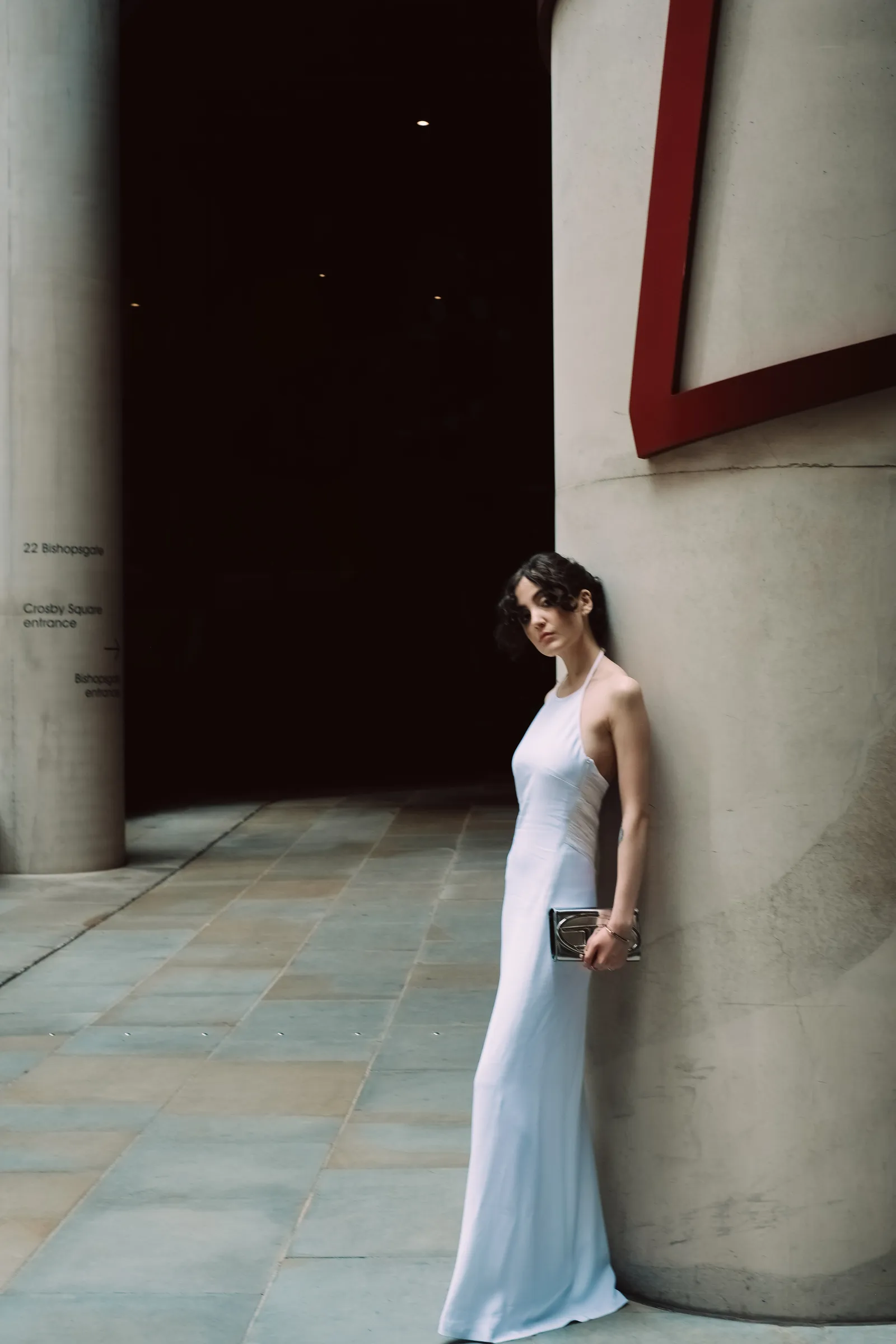 Woman in a pale gown leaning against a concrete column in a dark passage