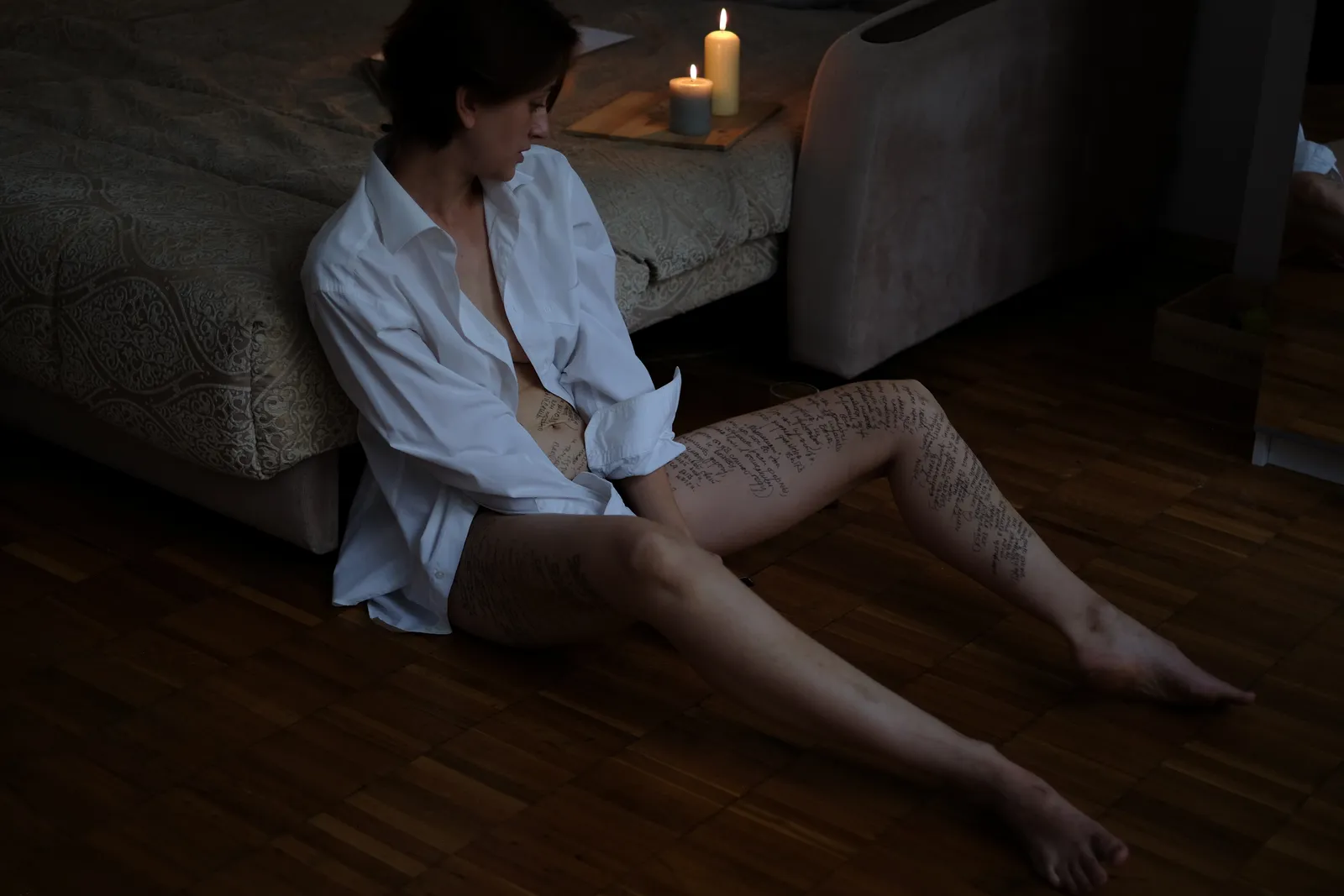 Woman in a white shirt on the floor against the bed, a candle above her, an outstretched leg covered in handwriting