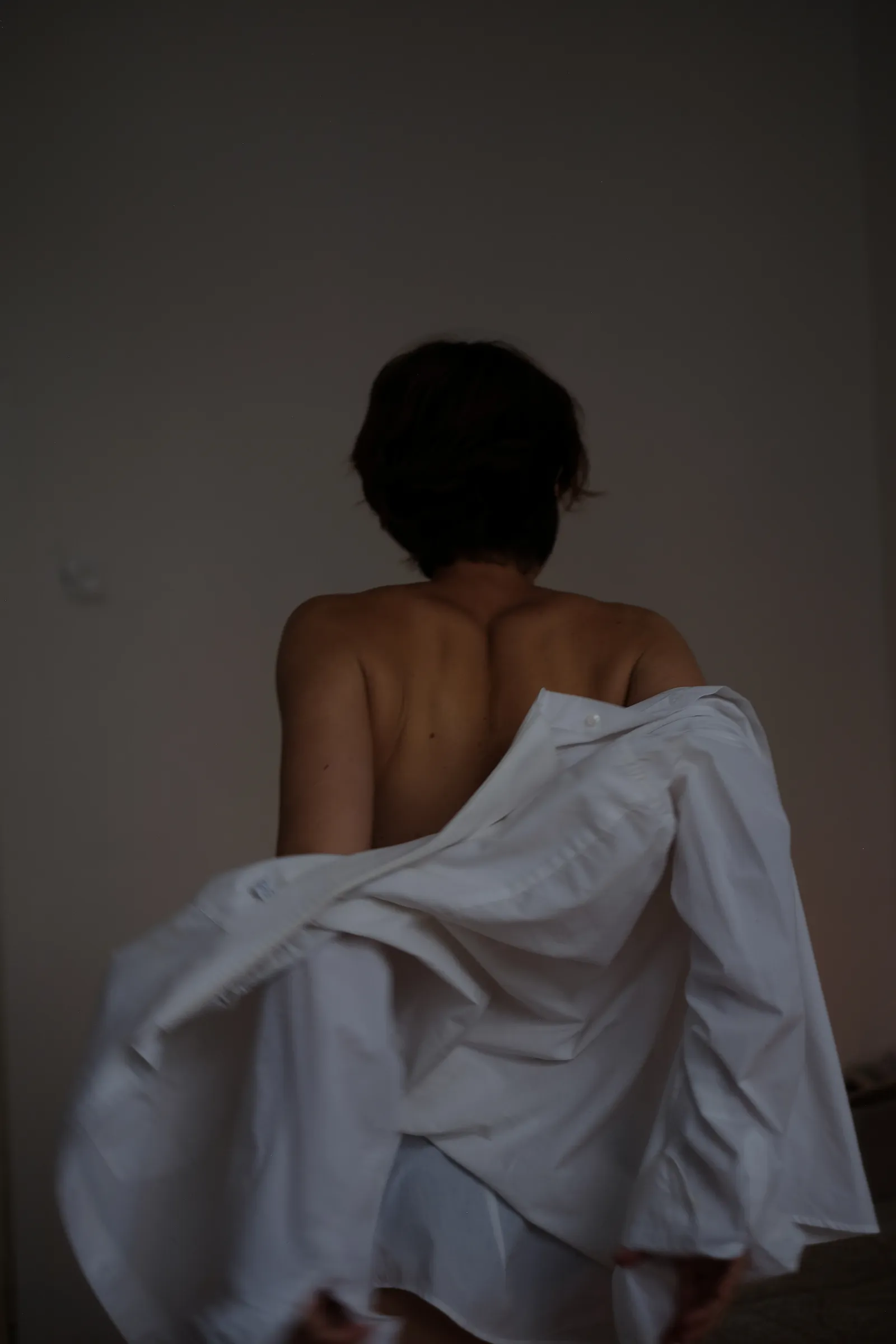 Bare back of a woman pulling a white shirt off her shoulders in a pale room