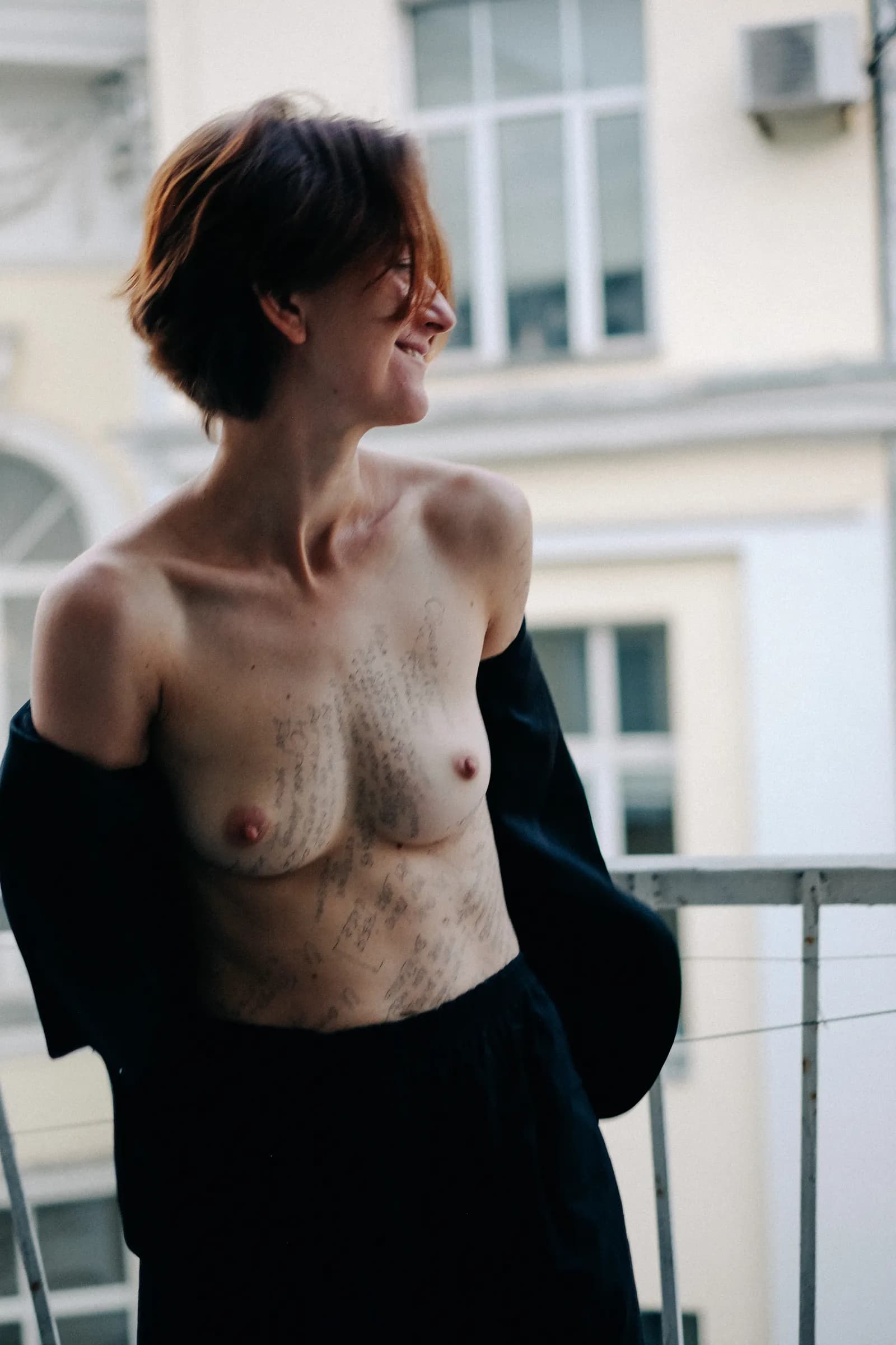Woman on a balcony smiling away from the camera, writing across her chest, pale facades behind