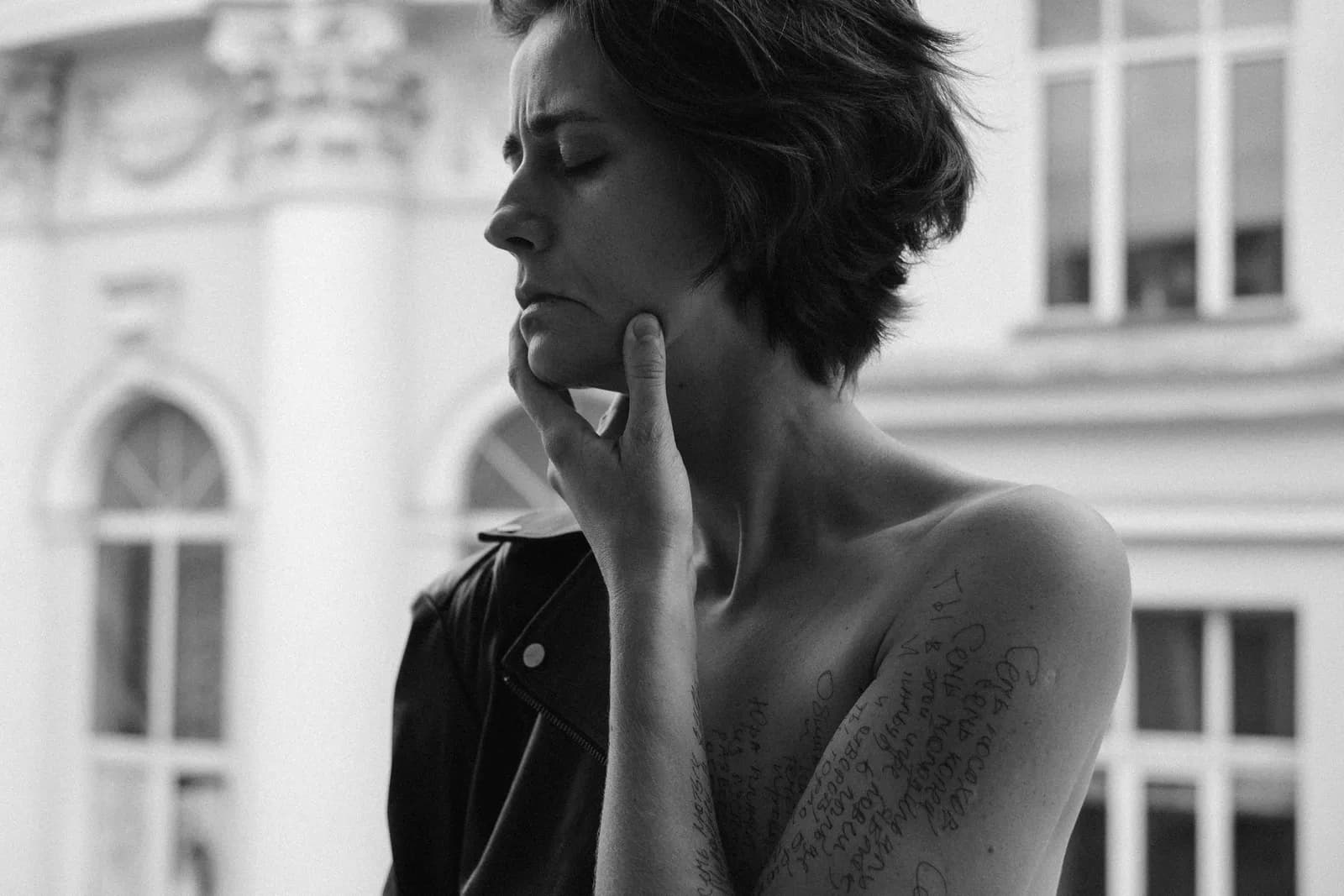 Black and white portrait of a short-haired woman on a balcony, hand at her chin, ink handwriting on her shoulder